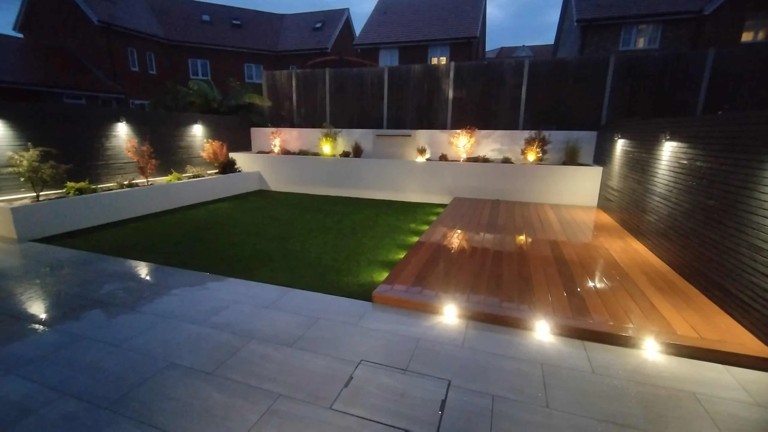 Modern garden with porcelain, hardwood decking and white rendered beds, lit at night